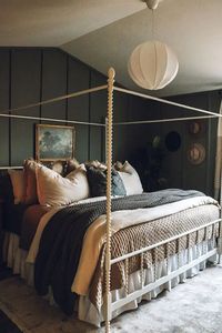 Picture of a moody attic bedroom with graphite grey walls, a white canopy bed with lots of neutral and dark pillows, a white pendant lamp and a hat gallery wall