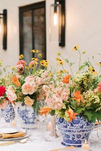 The arrangement includes a mix of cheerful yellow daisies, peach roses, orange lilies, and lush green foliage. The colorful blooms bring an air of freshness and warmth, perfect for an outdoor summer celebration or intimate dinner party. #SummerFlorals #TableSetting #OutdoorDinnerParty #FloralArrangements #ElegantTableDecor #SummerCelebration #VibrantFlowers #BlueAndWhiteVases