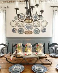 A vintage farmhouse dining room with paneled walls, a stained table, black chairs, a vintage plate wall and a chic chandelier