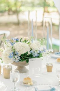 A small white floral centerpiece with blue accents. Included are blue taper candles to accent the blue in the arrangement and tea lights in gold votives. All florals by Vintage Soul Floral Design.