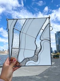 A minimalistic map of NYC made with Bullseye Glass. The glass used for the land (Manhattan, Queens, Brooklyn, and NJ) is a glossy opaque white and the glass used for the Hudson and East River is a textured iridescent clear.  Details: * Made with lead-free solder. Touch as much as you'd like! * Measures 10.5" long and 9.5" wide.  * Hangs 13.5". I suggest hanging from two S hooks from a curtain rod or from two screw nail hooks in a window seal.  * DO NOT hang with a suction cup hook. It might fall