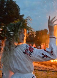 A girl is dressed in a traditional outfit, adorned with floral patterns, and wears a wreath made of greenery and flowers on their head. They are positioned near a bonfire with other people and trees visible in the background.