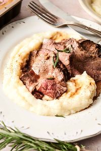 Slices of roasted beef atop a bed of mashed potatoes, garnished with rosemary, on a white plate with a fork beside it.