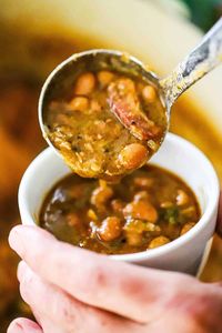 A person using a ladle to transfer a helping of borracho beans into a small white cup filled with the beans.