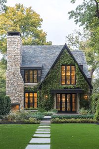 Modern european cottage house with stone siding chimney multi pitched gabled roof with slate tile modern windows with black trim vines on facade. Embrace the cozy charm and character of European cottages, where every stone and beam tells a story worth hearing.