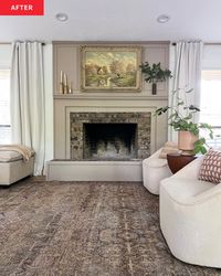 Fireplace after DIY makeover. Brick has been mostly painted over with warm gray paint, a mantel has been added. Mantel has candles, a plant, and there's art in a gold frame over the mantel. White curtains on windows to either side, 2 white chairs with brown patterned rug