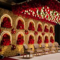 A stunning and opulent scene of an indoor space meticulously decorated for a grand Indian wedding or formal celebration. The wall is adorned with multiple arches, each housing exquisite red floral arrangements. A lavish canopy of red roses and white flowers hangs from the ceiling, illuminating the space with numerous hanging lights. Elegant upholstered chairs with intricate designs add to the regal and sophisticated atmosphere. The rich color palette of red and gold creates a luxurious and sumptuous environment, perfect for a night of unforgettable festivities.