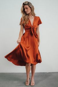 A fun twist on a classic satin dress Glowing rust color Satin material that reflects light beautifully Fit runs small Front tie at bust with peek-a-boo gap Short sleeves with subtle flutter Smocked at back waist Fully lined excluding sleeves High-low midi skirt Bump and nursing friendly 100% Polyester Hand wash cold, hang to dry, do not bleach We recommend steaming to release any wrinkles Skye is 5'9, cup size 32A, size 0 and wearing size S Anastasia is 5'4, cup size 38DD, size 14, and wearing X