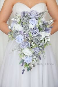 This lovely assortment of dusty lavender, lavender and periwinkle roses is bound to turn heads on your wedding day. This lovely cascading brides’ bouquet from the is an absolutely breathtaking arrangement for any bride to be, in any season. Silver glitter accents complete the look and add a delicate realism to this beautiful bouquet. This bouquet is 10” wide, 18” long, and is crafted by our in-house florists without using a foam base to anchor the flowers to the bouquet. Instead, we wire our bou