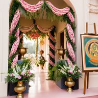 a luxurious and elaborate floral arrangement, possibly part of a wedding or a ceremonial event. The setup includes hanging floral chandeliers, large flower sculptures resembling lotus flowers, and intricately designed floral arches. The backdrop includes lush greenery, and the setting appears to be outdoors or in a large open space with a high ceiling. The overall ambiance is elegant and opulent, with a strong emphasis on floral decor.