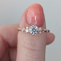 This delightfully whimsical engagement ring features 5 scattered side stones on each side of the center stone, reminiscent of glimmering stars scattered across the night sky. Details: Center Stone Specifications: Center Stone: Moissanite Colour: DEF (colourless) Shape: Round Brilliant, Hearts and Arrows Cut Measurement: Approximately 5.5mm D.E.W: Approximately 0.66ct Side Stone Specifications: Moissanite Colour: DEF Shape: Round Measurements: 1mm-1.3mm Ring Measurements: Band Width: Approx 1.8mm