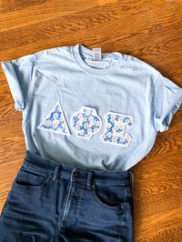 These classic and vintage-style letters are perfect for new and seasoned sorority women alike! Features double-stitched cottage-style blue daisy letters on a stark white background attached to a comfortable unisex baby blue tee.  This seller is licensed with Affinity Licensing to ensure a verified, quality product.    Care Instructions: Machine wash cold, inside out; Tumble dry low; Low iron as needed