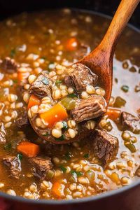 A close-up shot of a wooden spoon scooping beef barley soup from a pot. The soup is rich and thick with chunks of tender beef, whole barley grains, diced carrots, and finely chopped herbs, showcasing a hearty and nutritious meal.