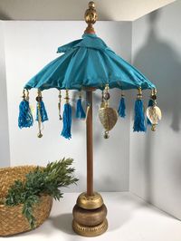 "Vintage Boho umbrella decor, Turquoise blue cloth decorative umbrella on wooden base with tassels trim, Bohemian style umbrella decor.  This umbrella is on a wooden base with a cloth blue canvas like umbrella with gold and blue tassel trim.  The umbrella opens and has a pin to hold it up, but there is no hole to put the pin in.  The umbrella will stay up without the pin as shown in photos but should probably have a hole drilled into the wood pole if wanted to keep it up permanently. Height 27\" Width 4.5\""