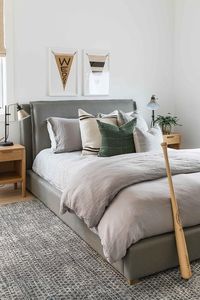 Cottage style boy's room features a dark gray upholstered bed on a gray jute rug, oak nightstands lit by black and brass lamps and framed vintage banners mounted over the bed.