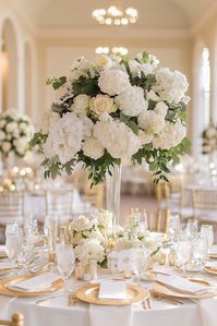 Crisp white hydrangeas and roses form this breathtaking tall wedding table centerpiece, lifted high on a clear vase for maximum impact. Surrounding the base with smaller arrangements and votive candles adds depth and soft light, creating a cohesive and sophisticated look for any venue.