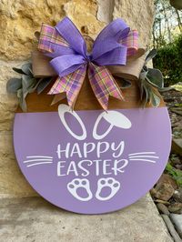 What a great way to welcome friends and family for the Easter holiday with this beautiful Happy Easter door hanger! This 18" round hanger features a stained base with lavender painted on bottom 3/4 and white lettering. Hanger is adorned with lambs ear and a beautiful multi layered bow. Please note that exact ribbons may not be available and a comparable combination could be provided.)  Please allow 1-2 weeks processing time for your order. Send a message if you would like to customize colors.  B