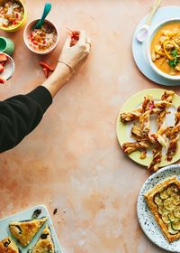 Blog post image showing a hand setting a food image scene by placing a piece of fruit near a bowl of food. On the edges of the iimage are plates with soup and various pastry creations.