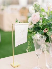 Small angled fabric table number on a tall gold Style Me Pretty stand. Off-white fabric with gold ink featuring elegant script and a modern block font table number for the Merrimon-Wynne House Winter Bridal Showcase in Raleigh, North Carolina. Photo by Alaina Ronquillo Photography.