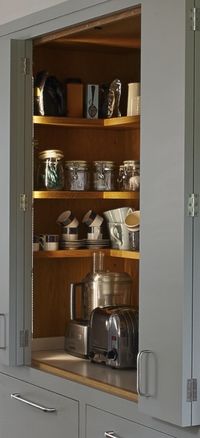 Grey bi-fold kitchen cupboard doors reveal wooden shelving inside a larder cupboard for food and appliance storage. Kitchen designed for Figura