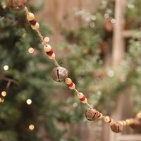 Bead and Bell Garland features rusty metal jingle bells and wooden beads in cream and red colors, all strung on a jute string garland with loops at each end for hanging. Bells measure 1" high and garland measures 5 foot long.