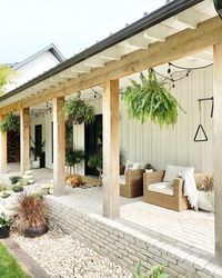 Board and batten siding is paired with wood columns attached to a wood beam. Woven chairs with white cushions sit upon a brick porch among a variety of greenery. Black strings of lights are draped throughout the ceiling.