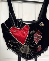 This soft black velvet boned corset features heavy embellished gold star and glittering sun details, along with embroidered hearts and a handmade padded heart embellished with lace, beads and features an embellished winking eye. The corset is filled with wonderful beading and sequin sequin designs making it extremely unique and certainly a stunning piece of artwork. This corset would be perfect for giving your valentines outfit the wow factor that you've been looking for! Corsets are lined with