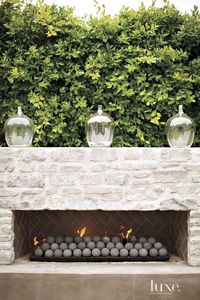 Chic patio features a pair of wicker sofas facing each other across from a weathered teak coffee table alongside weathered teak end tables placed in front of a white stone fireplace accented with a taupe herringbone firebox topped with hand blown jars.