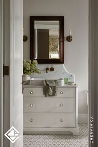 Old-world charm meets modern flair in this guest bathroom. ✨ Every detail shines, from the chic reeded drawers and charming knobs to the soft eased edges and scalloped furniture-style baseboard with a toe kick. Sealed in a warm white and crowned with a curved marble backsplash and an adorable wall-mounted faucet, this space is the perfect blend of elegance and charm...#BathroomDesign #BathroomVanity #FlutedVanity #FlutedDrawers #BathroomRemodel #BathroomIdeas