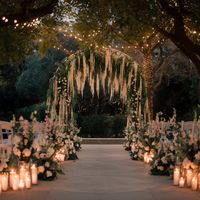 A photo of a mesmerizing outdoor wedding scene within a charming garden gazebo. The bride, radiant in a beautifully detailed white gown, walks gracefully down a flower-lined path, escorted by her father. The ambiance is tranquil and refined, with a profusion of floral arrangements and lush greenery. The officiants and groom stand poised at the end of the path, eagerly awaiting the bride's arrival. The scene artfully combines natural beauty, romance, and elegance, capturing the essence of a perfect, love-filled wedding day.