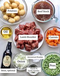 All recipe ingredients on a white board, labeled in separate bowls: Potatoes, beef stock, oil, lamb shoulder, carrots, stout, onion, arrowroot, peas, thyme and garlic powder.