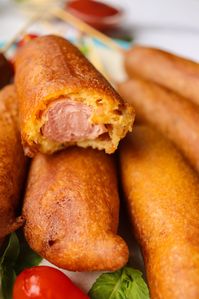 Close-up of several corn dogs, with one partially eaten, revealing the sausage inside. The corndogs are arranged on a white surface with green leaves and a red pepper in the background, reminiscent of a State Fair treat.