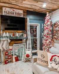 A skinny tree bursting with red and white ornaments is placed in the corner of this rustic farmhouse living room. A gray couch is seated next to the tree in front of a wooden faux fireplace. White, red, and tan Christmas décor arranged around the fireplace proves more is merrier.