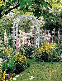 Paint can add a style boost to a simple arbor without overwhelming a vibrant garden setting. This rustic arbor stands in contrast to a riotous display of colorful flowers thanks to a coat of white paint. The pretty arched design creates a smooth transition between different areas of the garden. #archtrellis #gardenarch #archwayideas #landscaping #gardenarbor #bhg