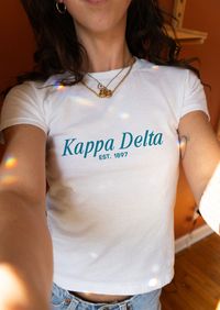 Introducing the Kappa Delta Baby Tee - a trendy and spirited addition to your sorority wardrobe! This adorable tee features a modern beachy-inspired design. Crafted from top-notch fabric, this crop tee guarantees both comfort and durability. Its soft and cozy interior provides a snug fit, making it an ideal choice for various occasions. Whether you're heading to a chapter meeting, attending social events, or spending quality time with your sorority sisters, this retro tee effortlessly complement