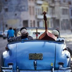 two people in an old blue car driving down the street with their backs turned to the camera