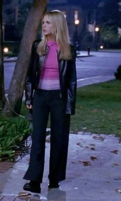 a woman standing on the sidewalk in front of a tree and wearing a black leather jacket