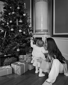 a woman holding a baby in front of a christmas tree with presents on the floor