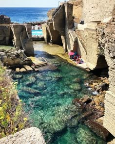 people are swimming in the clear blue water next to an old stone wall and bridge