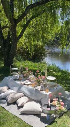 an outdoor table is set up with flowers and plates on it, near the water