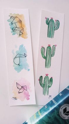 two watercolor bookmarks with drawings of cactuses and paper planes on them next to a bookshelf