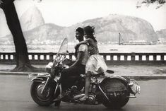 two people riding on the back of a motorcycle down a street next to a lake