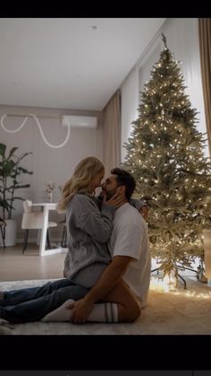 a man and woman sitting on the floor in front of a christmas tree with lights