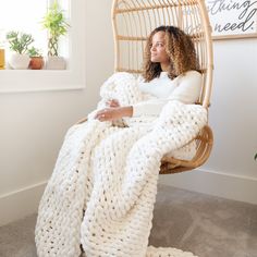 a woman sitting in a wicker hanging chair under a white knitted blanket on the floor