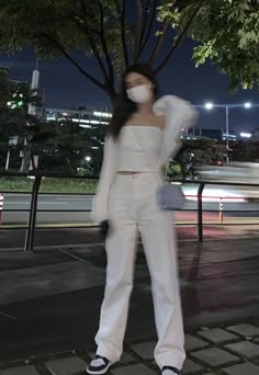 a woman in white is standing on the sidewalk with her hands behind her head and wearing a face mask