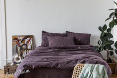 a bed with purple sheets and pillows next to a potted plant in a white room