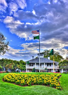 a flag flying in front of a white house with yellow flowers and green grass on the lawn