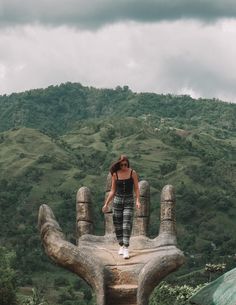 a woman standing on top of a giant hand statue in front of a lush green hillside