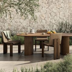 a wooden table with two chairs around it near a stone wall and some plants on the ground