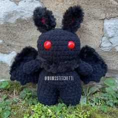 a black crocheted stuffed animal with red eyes and ears sitting in front of a stone wall